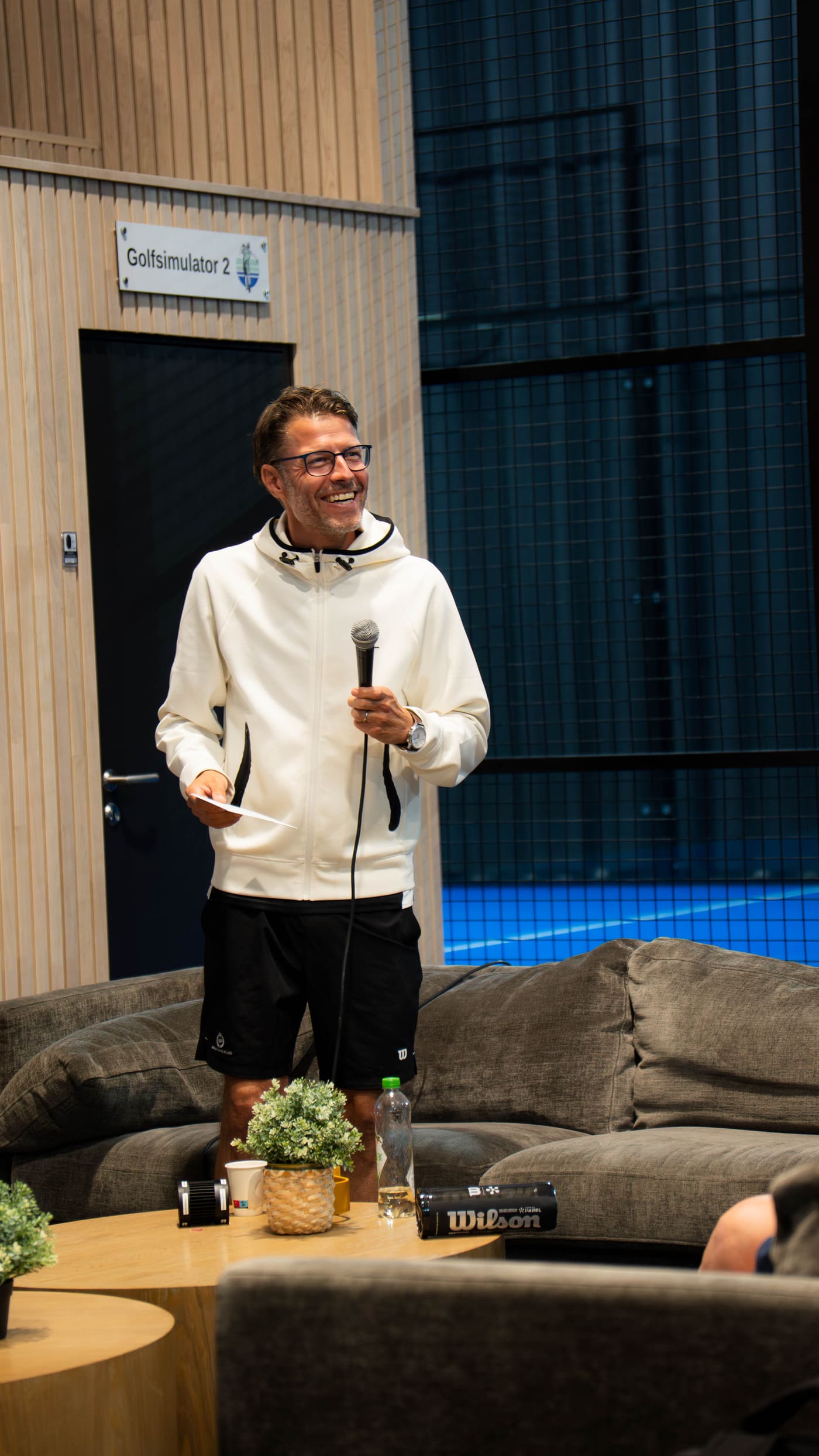 Mann i hvit hettegenser holder mikrofon foran padelbanen