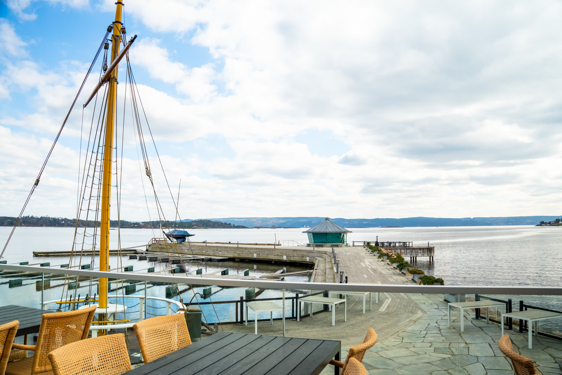 Utsikt over brygga med seilbåtmast og glasspaviljong ved fjorden
